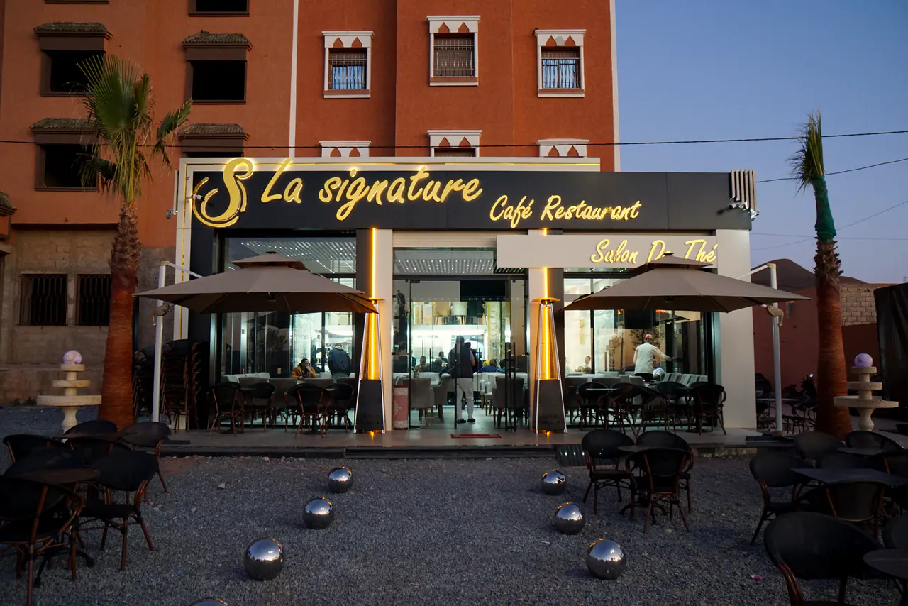 La façade de La Signature à l’heure bleue : enseigne dorée, terrasse et palmiers
